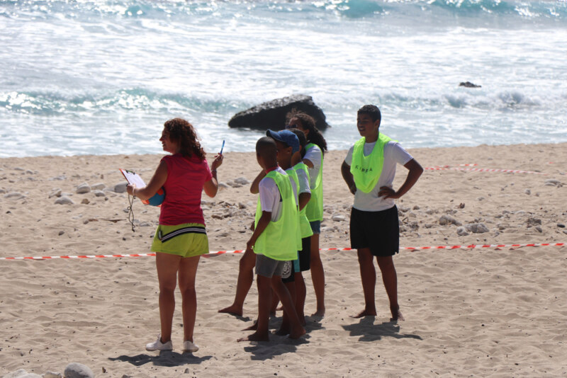 Groupe d’adolescents portant des chasubles fluorescentes écoutant les consignes d’une animatrice sur une plage, face à l’océan